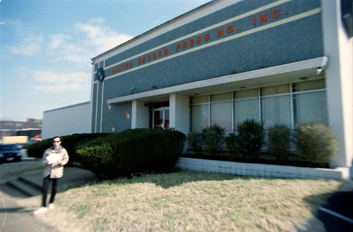A person walks by the United Record Pressing building, featuring a gray facade and large windows.