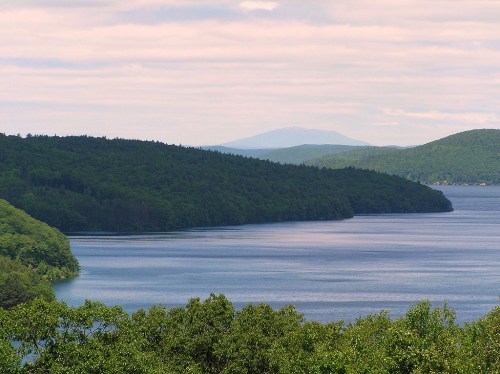 A serene lake surrounded by lush green hills under a cloudy sky, with distant mountains in the background.