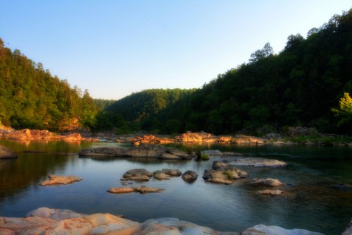 A serene river scene with smooth rocks, surrounded by lush green hills under a clear blue sky.