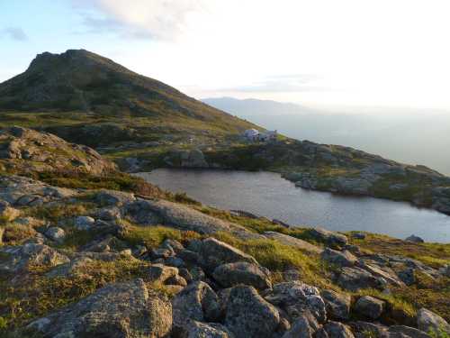 A scenic mountain landscape with rocky terrain, a small lake, and a distant building under a soft sunset sky.