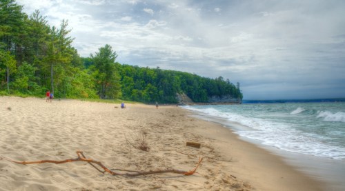 A sandy beach with gentle waves, surrounded by green trees and cliffs under a cloudy sky. People stroll along the shore.
