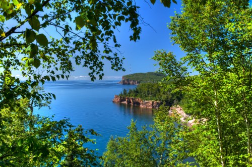 A serene coastal view framed by lush green trees, showcasing calm blue waters and distant cliffs under a clear sky.