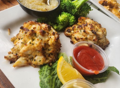 Two crab cakes served with lemon wedges, lettuce, broccoli, and dipping sauces on a white plate.