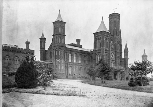Historic black-and-white photo of a large, ornate building with towers and a clock, surrounded by trees and a pathway.