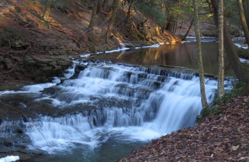 A serene waterfall cascades over rocks into a calm stream, surrounded by trees and autumn leaves.