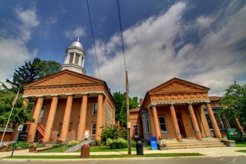Historic brick building with columns, a dome, and a library entrance, set against a cloudy sky. Greenery surrounds the area.