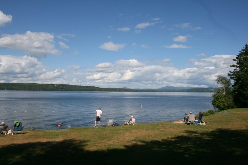 A serene lakeside scene with people relaxing on the shore, surrounded by trees and a blue sky with fluffy clouds.