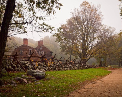 A foggy scene featuring a rustic house behind a stone fence, surrounded by trees with autumn foliage.