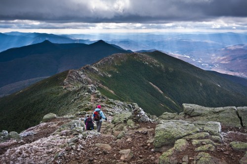 Two hikers navigate a rocky mountain trail, surrounded by dramatic clouds and distant peaks under a moody sky.