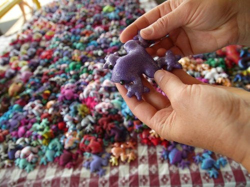A person holds a colorful, textured frog figurine above a large collection of similar figurines on a patterned surface.