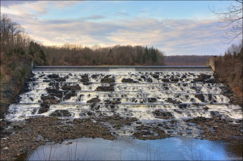 A cascading waterfall flows over a dam, surrounded by trees and a cloudy sky. Rocks are visible in the foreground.