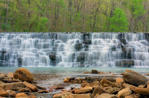 A serene waterfall cascades over rocks, surrounded by lush green trees and a calm riverbed.
