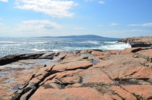 Rocky coastline with orange-hued stones, ocean waves, and distant hills under a clear blue sky.