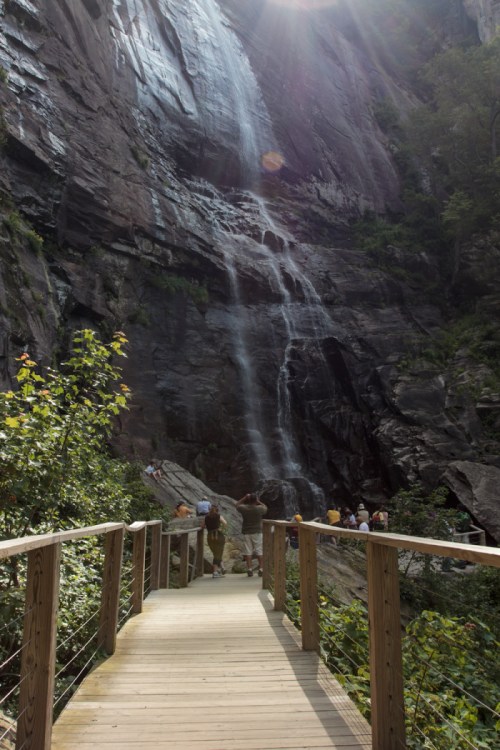 A wooden walkway leads to a waterfall cascading down a rocky cliff, surrounded by lush greenery and visitors.