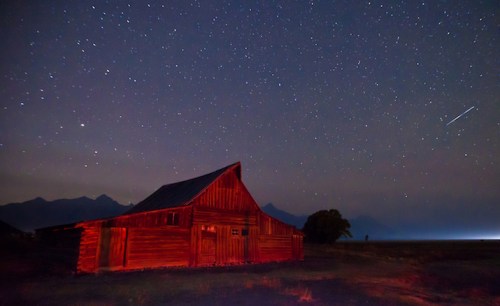 A rustic red barn under a starry night sky, with mountains in the background and a faint light on the horizon.