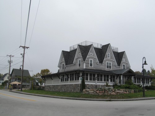 A large, multi-gabled house with a gray exterior, surrounded by greenery, on a quiet street under a cloudy sky.
