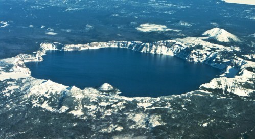 Aerial view of a large, snow-capped crater lake surrounded by mountains and forested land.