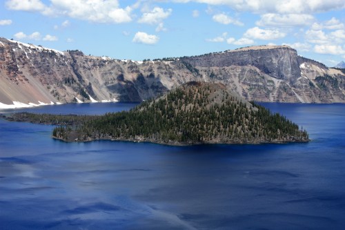 A serene view of a blue lake with a small island surrounded by mountains and trees under a partly cloudy sky.