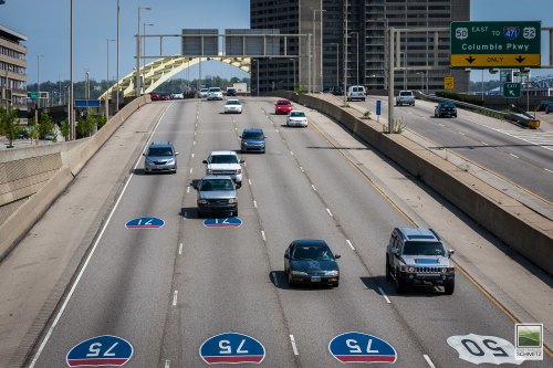 A busy highway with cars traveling, featuring road signs and a bridge in the background under a clear blue sky.