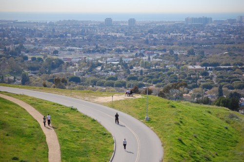 A scenic view of a winding road with people walking, overlooking a city and coastline in the distance.