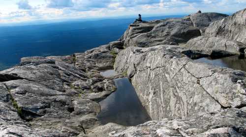 A person sits on rocky terrain with pools of water, overlooking a vast landscape under a cloudy sky.