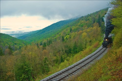 A steam train travels along a winding track through lush green mountains under a cloudy sky.