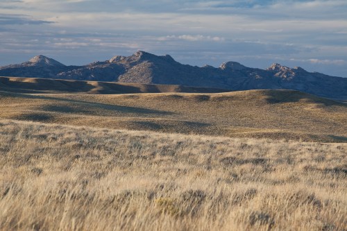 A vast landscape of rolling hills covered in grass, with rugged mountains in the background under a cloudy sky.
