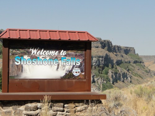Sign welcoming visitors to Shoshone Falls, with a scenic view of cliffs and landscape in the background.