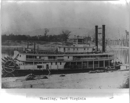 A historic steamboat docked in Wheeling, West Virginia, with two smokestacks and a scenic background.