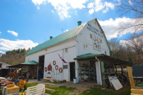 A white barn with a green roof, featuring a welcome sign and various outdoor decorations, set against a blue sky.