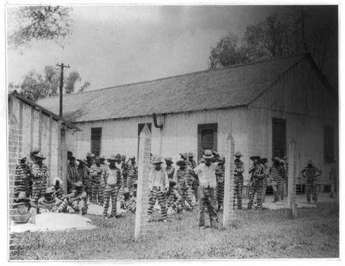 Historical black-and-white photo of a group of prisoners in striped uniforms outside a wooden building.
