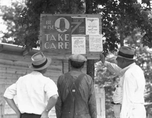Four men in hats examine a wooden sign on a tree, reading "Be Wise, Take Care" with various notices attached.