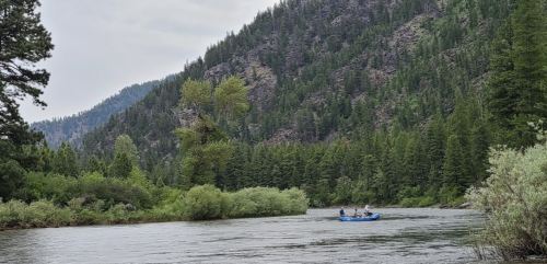 A blue raft with three people floats on a river surrounded by lush green trees and mountains in the background.