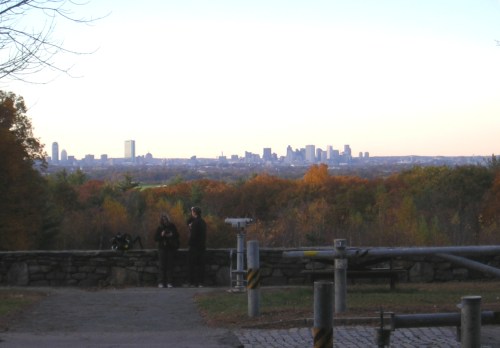A scenic view of a city skyline in the distance, framed by autumn trees and two people standing near a stone wall.