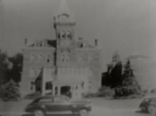 Historic building with a clock tower, surrounded by trees and vintage cars in black and white.