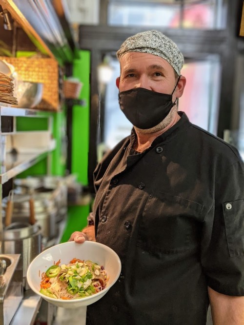 A chef in a black uniform and mask holds a bowl of food in a restaurant kitchen, surrounded by cooking equipment.
