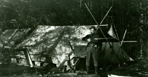 A man stands with a rifle in front of a tent, with a sign reading "Seldom Tim" nearby, in a wooded area.
