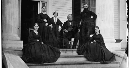 A historical black-and-white photo of six people in 19th-century attire, posed on the steps of a large house.