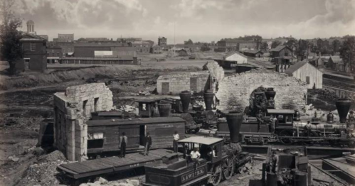 Historic black-and-white photo of a train yard with steam locomotives and ruins of buildings in the background.