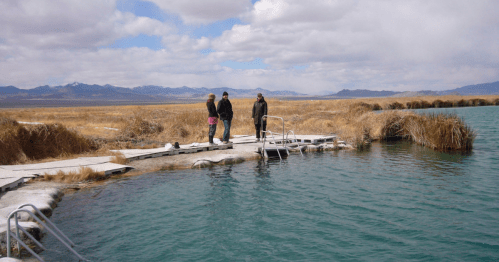Three people walk along a wooden dock by a turquoise hot spring, surrounded by dry grass and mountains under a cloudy sky.