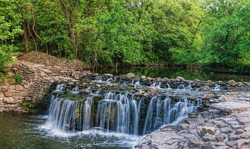 A serene waterfall cascades over stone steps, surrounded by lush green trees and a calm river.