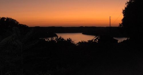 A serene sunset over a calm lake, with silhouettes of trees and hills against a vibrant orange sky.