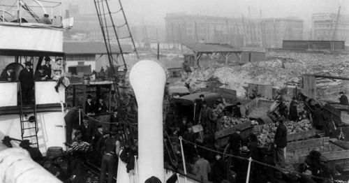 Black and white photo of a busy port with people, ships, and cargo in a foggy urban setting.