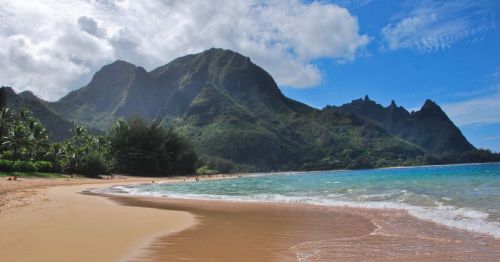 A serene beach with golden sand, clear blue water, and lush green mountains under a partly cloudy sky.