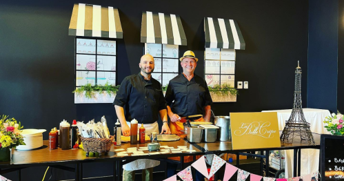 Two chefs stand behind a table with condiments and cooking supplies, decorated with flowers and a sign featuring the Eiffel Tower.
