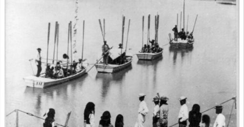 Black and white photo of boats on a river, with people fishing and standing on the shore.