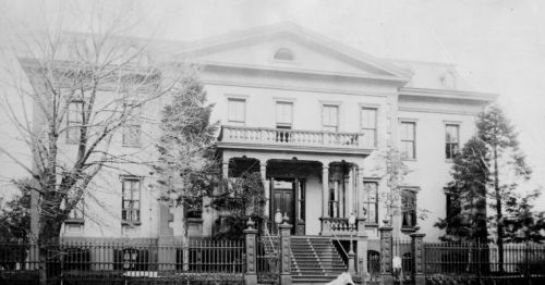 Historic black-and-white photo of a grand mansion with a large staircase and ornate balcony, surrounded by trees.