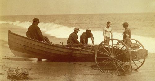 Five men on a beach near a wooden boat, with one man standing shirtless beside a cart filled with fishing gear.