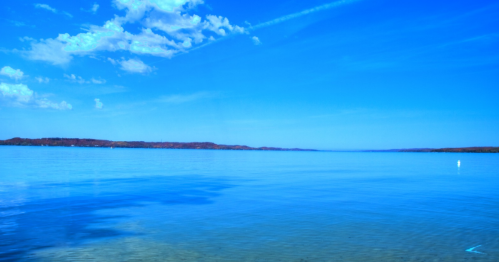 A serene blue lake under a clear sky, with distant hills and soft clouds reflecting on the water's surface.