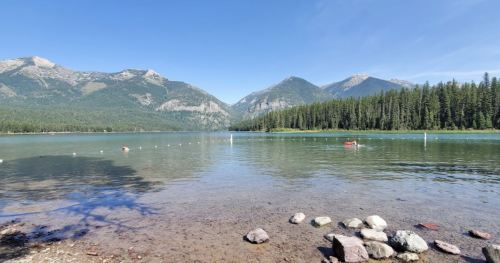 A serene lake surrounded by mountains and trees, with clear water and a rocky shore under a blue sky.